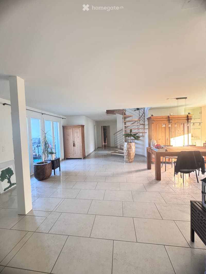 Bright, open corridor with tiled flooring, large windows, a wooden staircase, a dining table with chairs, and potted plants.