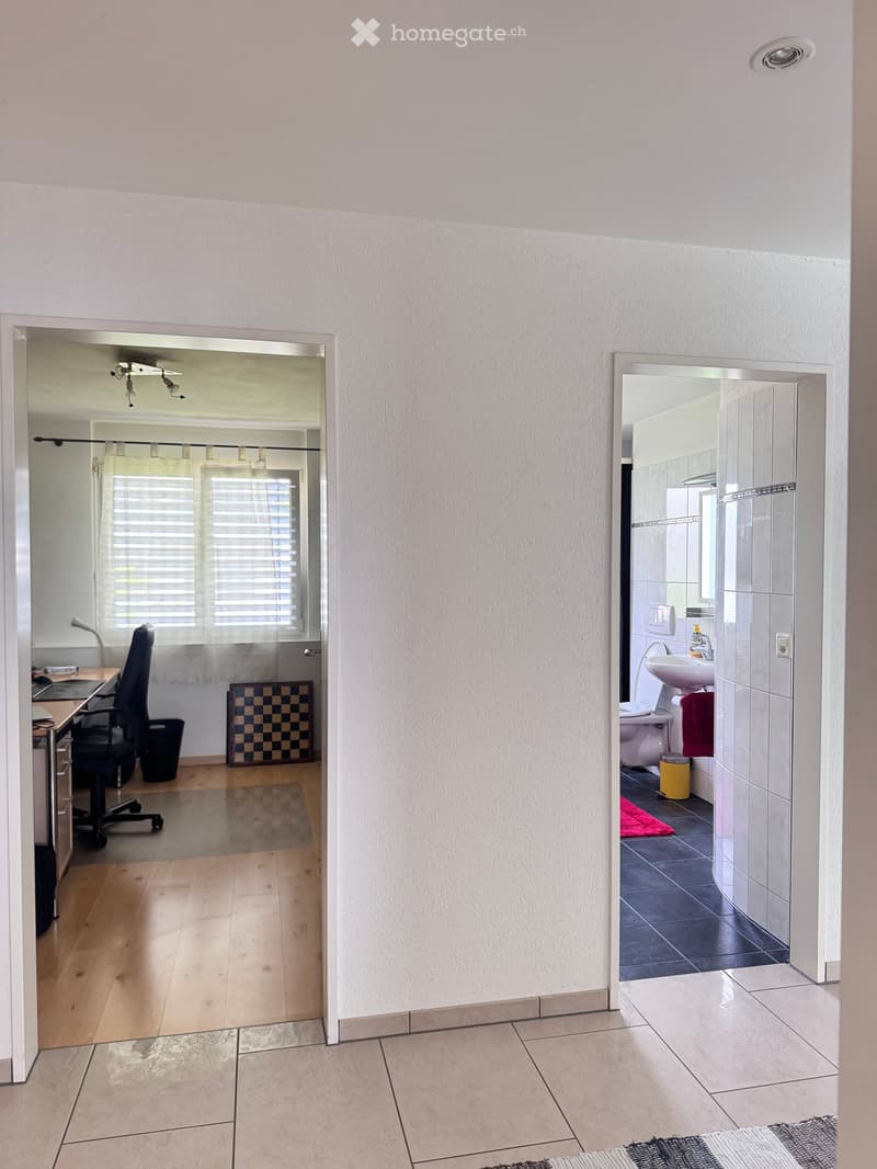 A hallway with light-colored walls and tiled floors, leading to a furnished room and a bathroom. The room contains a desk, chair, and a small rug, while the bathroom has a sink, toilet, and a red towel.