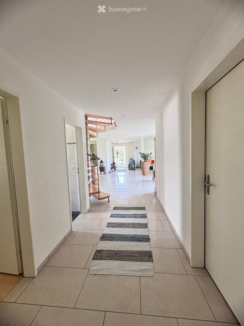 A long, well-lit hallway with tiled flooring, white walls, and a striped runner rug. A wooden staircase is visible to the left, leading to another level.
