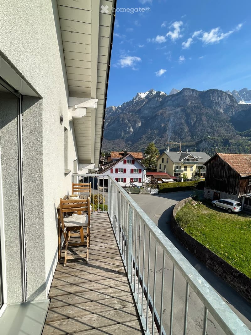 Balcony with wooden floor, metal railing, and a wooden chair, overlooking a scenic mountain view and a small town below.
