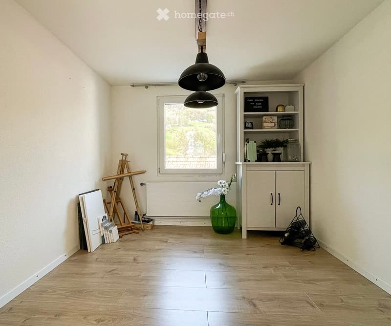 Bright room with wooden flooring, large window, hanging pendant lights, cabinet with decorative items, green vase with flowers, easel, and books stacked against the wall.