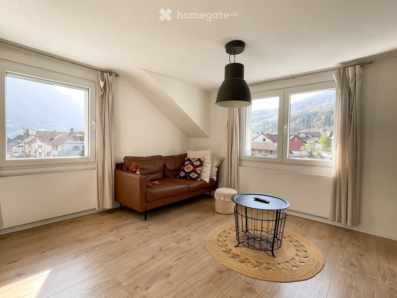 Living room with wooden flooring, large windows offering a scenic view, brown leather sofa, small round table, and decorative rug.