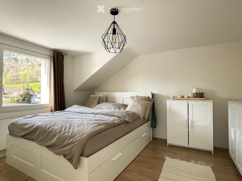 Cozy attic bedroom with a white bed, pillows, a hanging geometric lamp, a white wardrobe, and a window with a view of greenery.