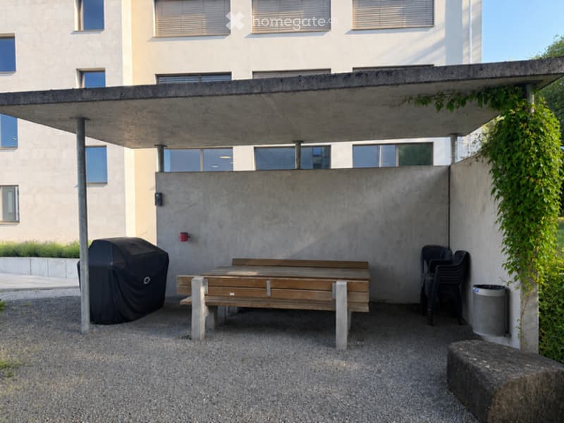 Modern outdoor seating area with a wooden bench, covered by a concrete roof, surrounded by a concrete wall and greenery. Includes a covered grill and plastic chairs.