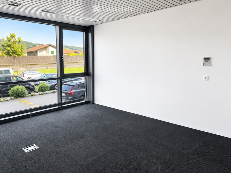 Empty room with white walls, grey carpet, large glass sliding doors, and a view of a parking lot and greenery.