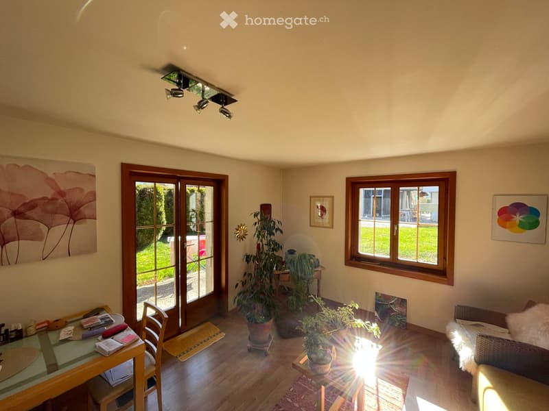 Bright living room with wooden flooring, large windows, and a sliding glass door leading to a garden. Features a dining table with chairs, a sofa, potted plants, and artwork on the walls.