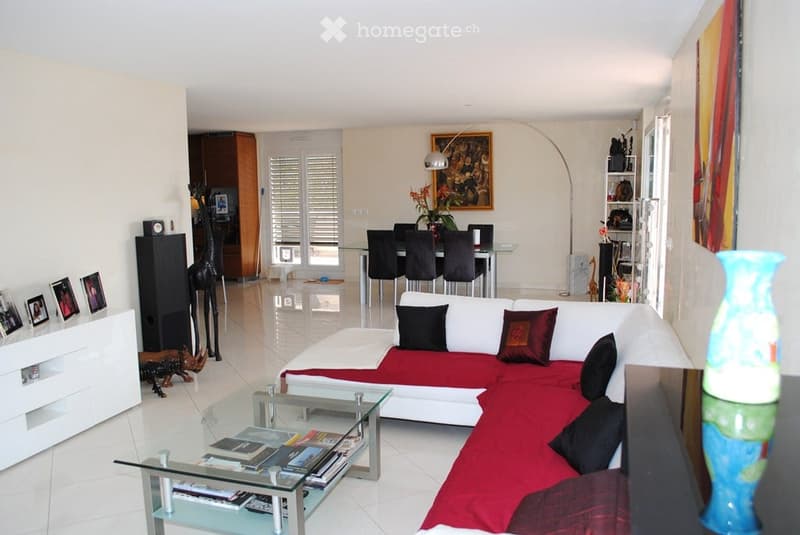 Modern living and dining area with white walls, tiled floor, white sectional sofa with red cushions, glass coffee table, dining table with chairs, floor lamp, decorative shelf, and artwork.