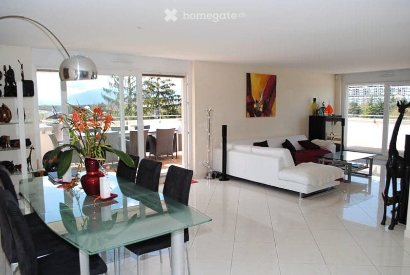 Bright, modern living and dining area with white walls, tiled flooring, large windows offering a scenic view, a white sectional sofa with an ottoman, a glass dining table with black chairs, a floor lamp, shelves with decorative items, and artwork on the w
