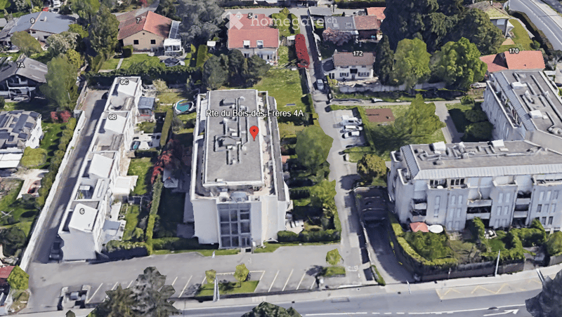 Aerial view of a multi-story residential building with a flat roof, surrounded by greenery and neighboring houses.
