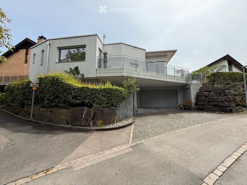 Modern two-story house with a flat roof, large windows, and a balcony on the upper level. The house features a garage and is surrounded by greenery and a stone wall.