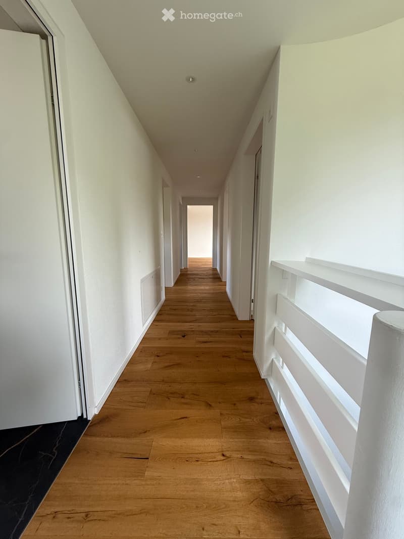 Clean, well-lit hallway with light wooden flooring, white walls, and white doors on both sides. A white railing is visible on the right side.