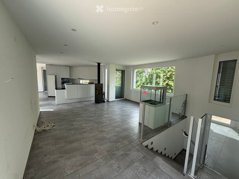 Empty room with tiled floor, white walls, and large windows. Features a staircase with glass railing and a visible kitchen area in the background.
