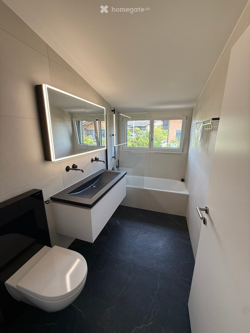 Modern bathroom with dark marble flooring, white walls, a sleek sink with a modern faucet, a bathtub, a toilet, and a large illuminated mirror above the sink. The room features large windows with a view of greenery outside.
