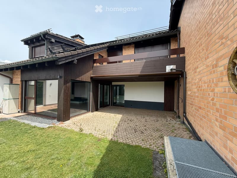 Courtyard with brick-paved pathways, grassy area, and modern building with wooden and brick facades, featuring a balcony and large windows.
