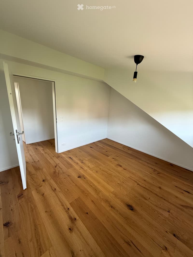 Empty room with wooden flooring, white walls, and a single hanging light fixture. An open doorway leads to another room.