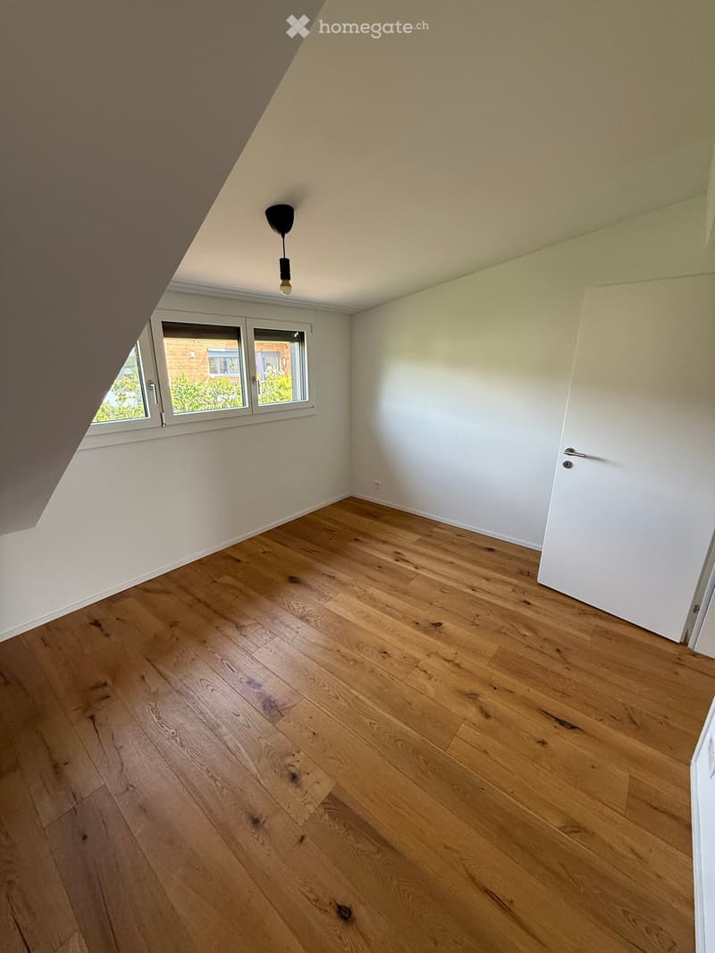 Empty attic room with wooden flooring, white walls, and a single window offering a view of the outside. The room features a hanging light fixture and a closed white door.