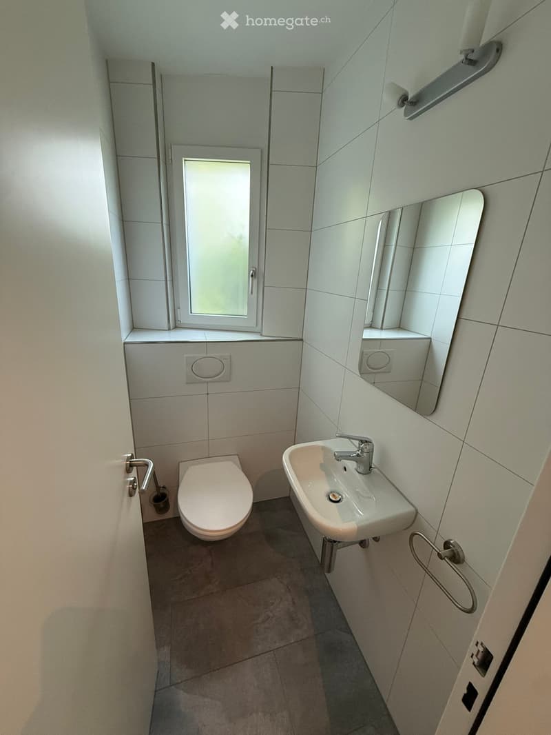 Modern bathroom with white tiled walls, a toilet, a sink with a faucet, and a large mirror above the sink. A small window provides natural light.