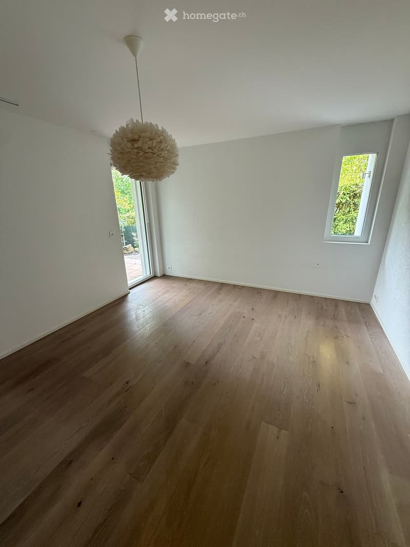 Empty room with light wooden flooring, white walls, and a large window with a view of greenery. A hanging decorative sphere is visible from the ceiling.