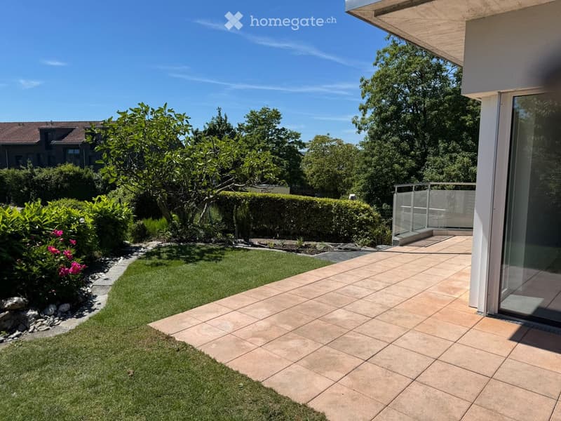 Well-maintained garden with a tiled patio, neatly trimmed grass, flowering shrubs, and a small tree, bordered by a low hedge and a stone pathway.