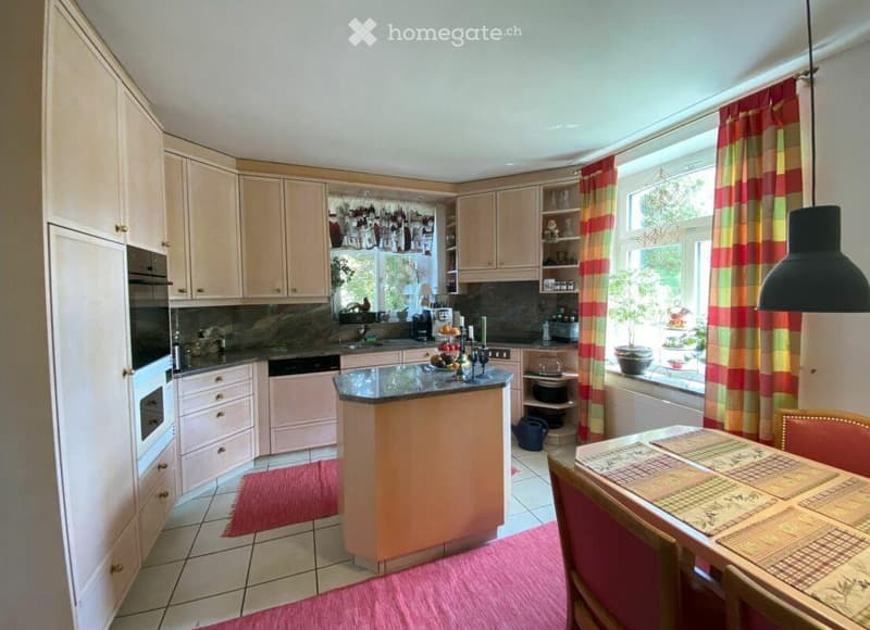 Bright kitchen with light-colored cabinets, a central island, and a dining area. Features include a sink, dishwasher, oven, refrigerator, and a large window with striped curtains.