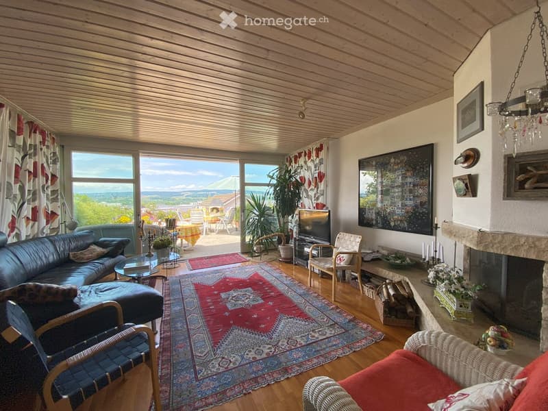 Bright living room with wooden ceiling, large windows offering a scenic view, a dark blue leather sofa, an ottoman, a wicker chair, a glass coffee table, a television, a fireplace with decorative candles, and a colorful rug.