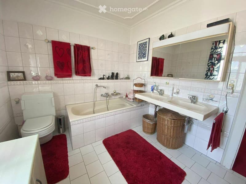 Bathroom with white tiles, a bathtub, toilet, double sink, mirror, red towels, and decorative items on the wall.
