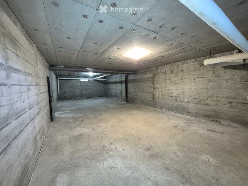 Empty underground parking space with concrete walls, ceiling, and floor. Features a single doorway and exposed pipes.