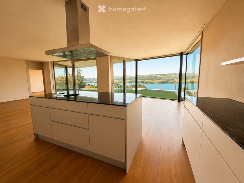 Modern kitchen with light-colored cabinets, a central island, and expansive windows offering a scenic view of a lake and landscape.