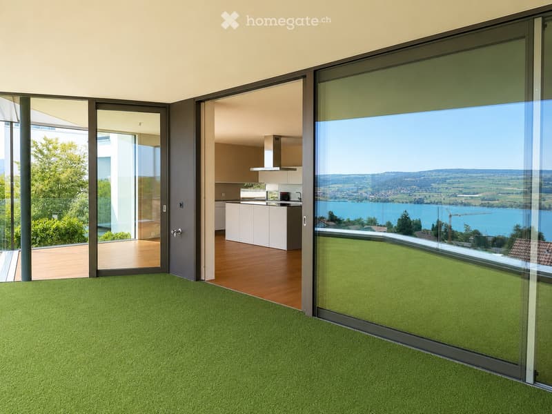 Bright, modern room with green carpeting, large glass sliding doors opening to a balcony, and a visible kitchen area with white cabinets and stainless steel appliances.