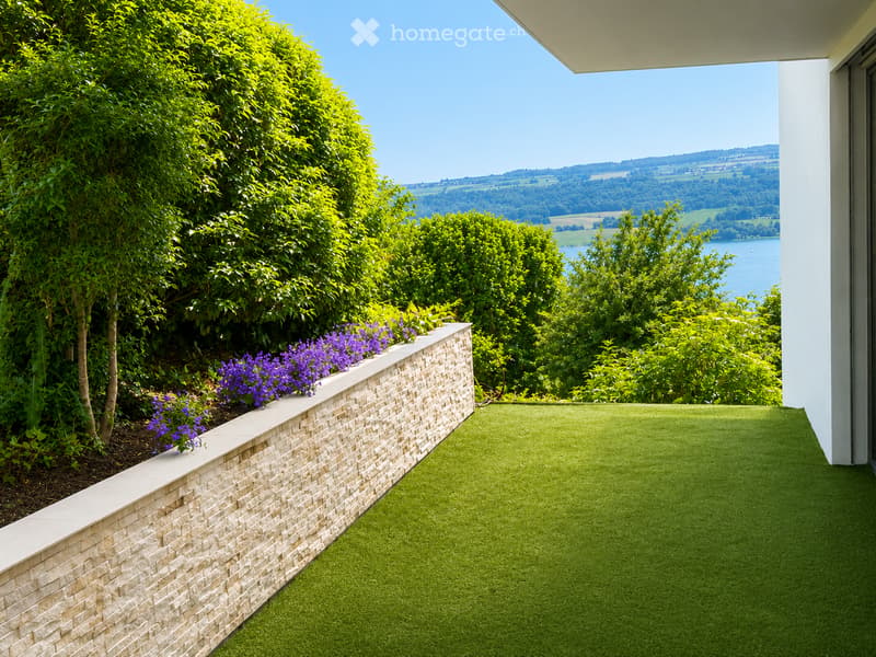 Balcony with artificial grass, stone low wall, purple flowers, and scenic countryside view.