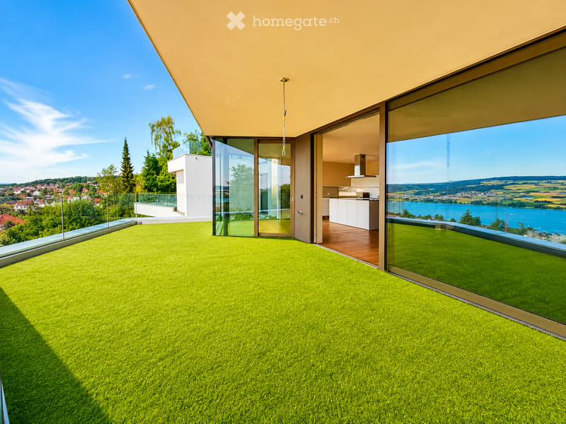 Modern balcony with artificial grass, large glass walls, and a view of a lake and surrounding landscape.