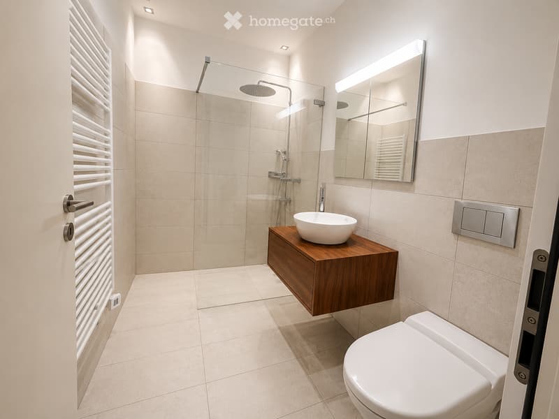 Modern bathroom with beige tiles, a white toilet, a wooden sink cabinet with a round sink, a large mirror with built-in lighting, and a glass-enclosed shower area with a rainfall showerhead.