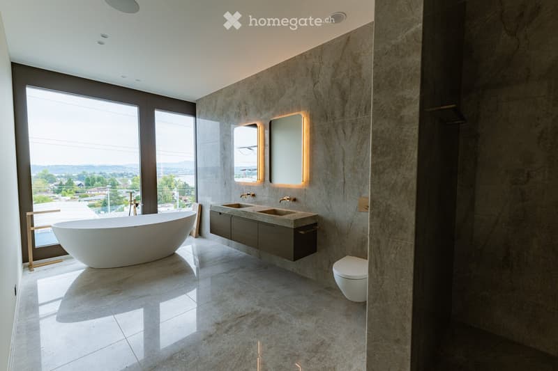 Modern bathroom with a large freestanding bathtub, double sink vanity, toilet, and mirrors with built-in lighting. Large windows offer a scenic view.