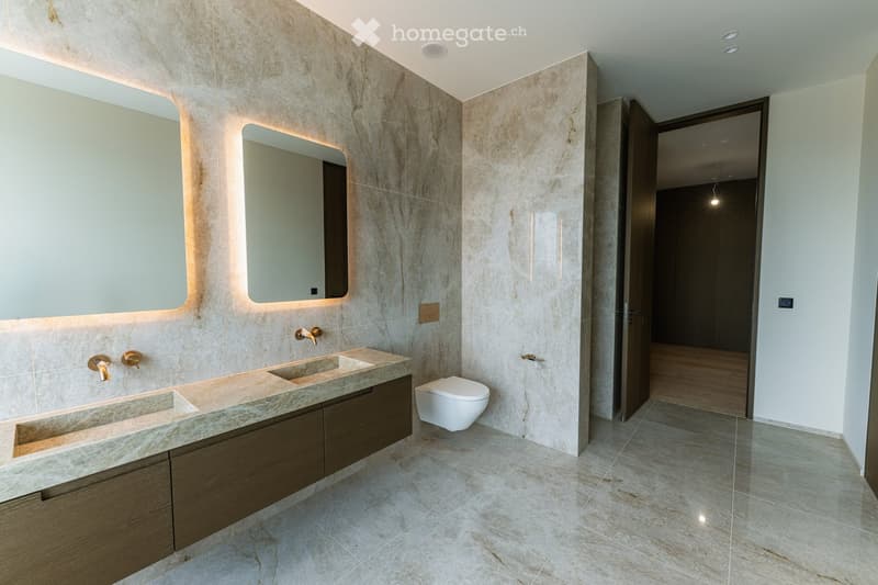 Modern bathroom with marble walls and floors, double sink vanity with wooden cabinets, illuminated mirrors, and a toilet.