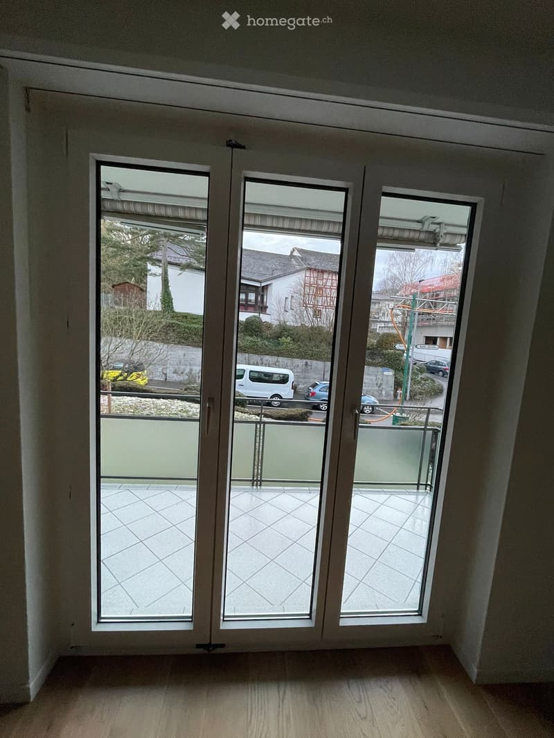 Empty room with wooden flooring and large sliding glass doors leading to a tiled balcony area. The balcony overlooks a street with parked cars and buildings.