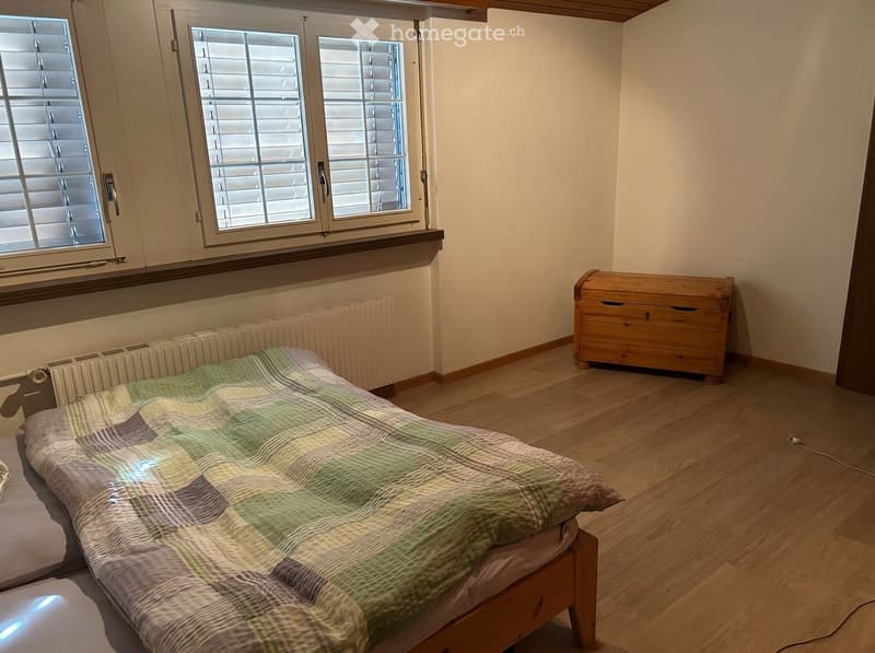 Bedroom with wooden floor, bed with green and brown quilt, wooden chest, radiator, and window with blinds.