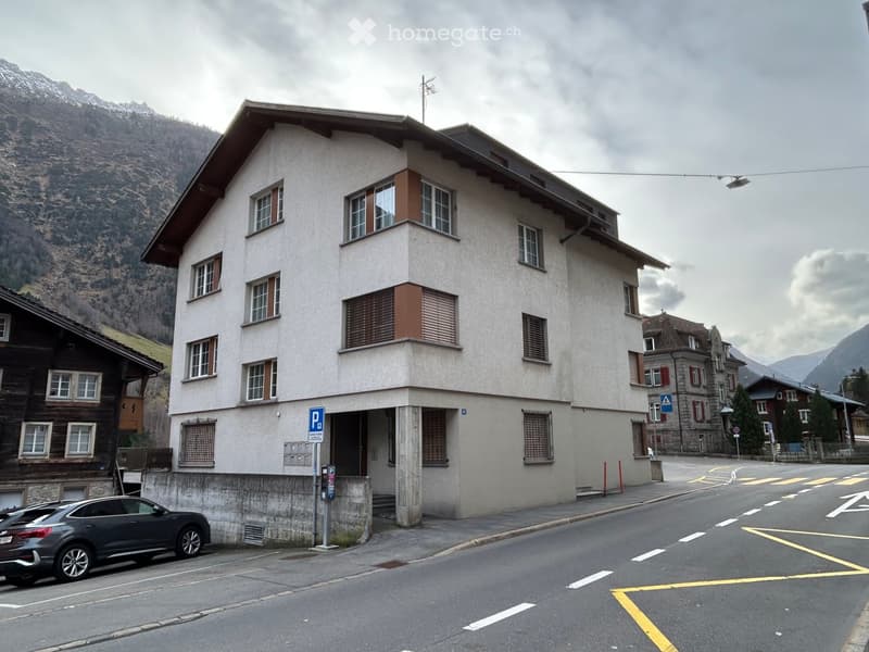 A multi-story building with a light-colored facade, multiple windows, and a small entrance area. The building is situated on a street with a parked car and a parking sign nearby. The surrounding area includes other buildings and a mountainous backdrop.