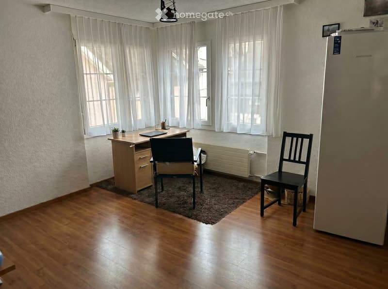 A furnished room with wooden flooring, a desk with a laptop, two chairs, a radiator, and a refrigerator. The room has large windows with white curtains and a small plant on the desk.