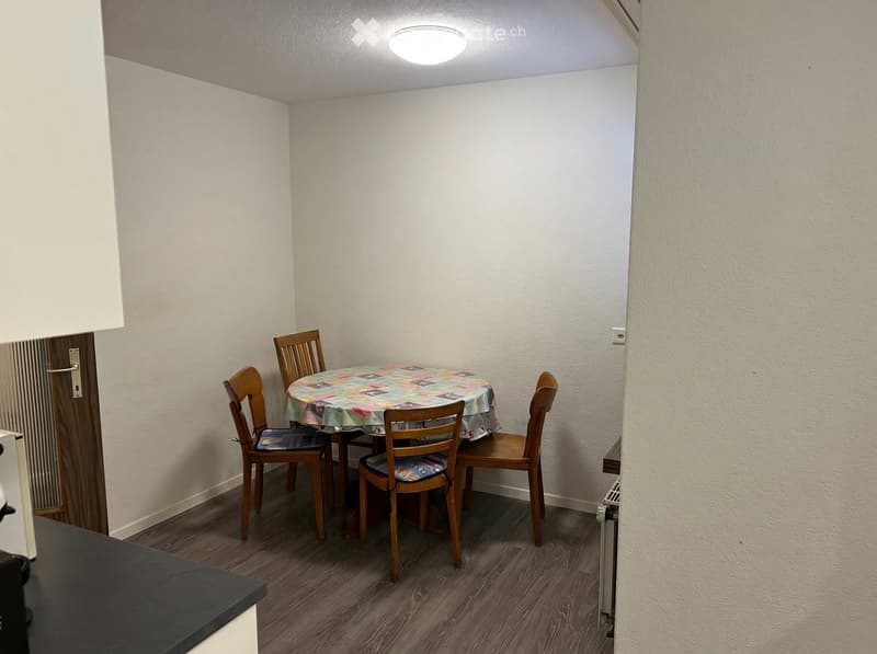 Small dining area with a round table covered in a colorful tablecloth and four wooden chairs, set against white walls and wooden flooring.