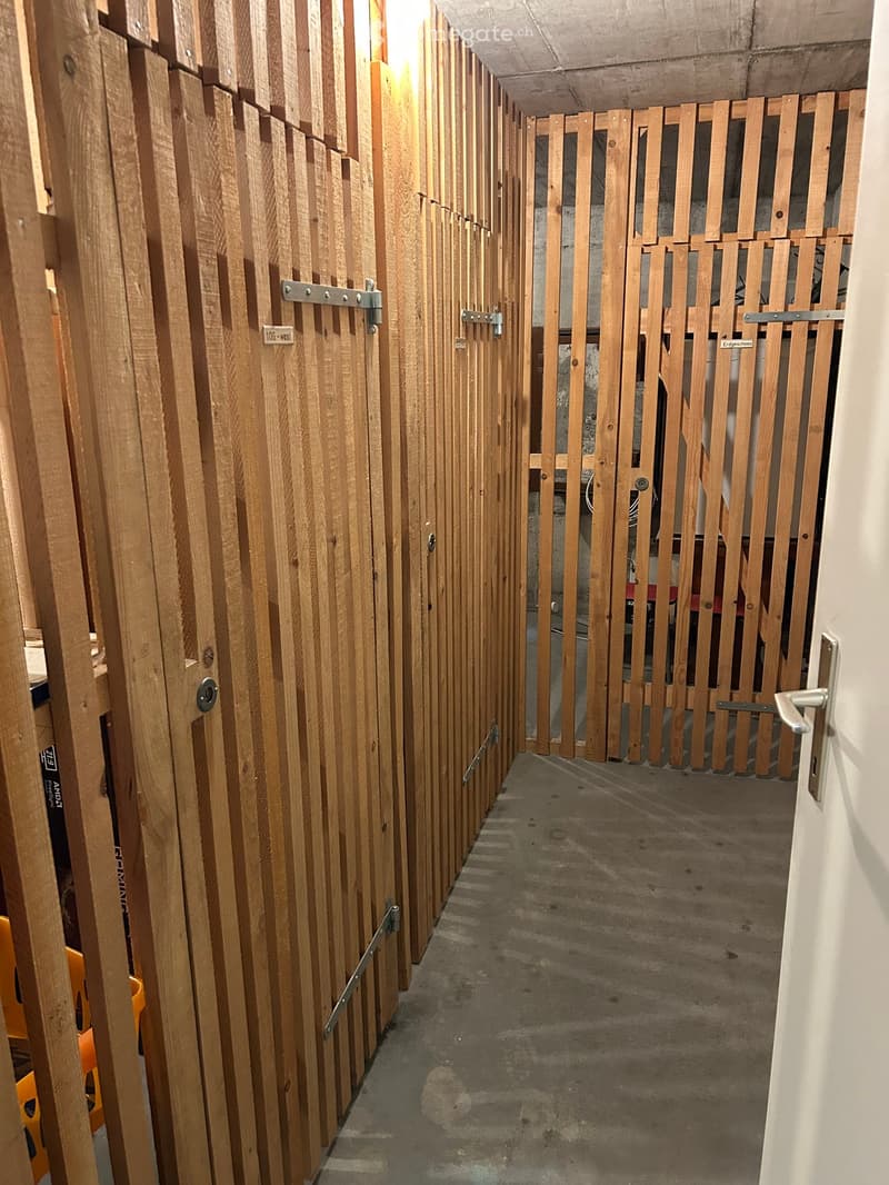 A narrow hallway with wooden panel walls and metal latches on doors, concrete floor, and a visible light fixture at the end.
