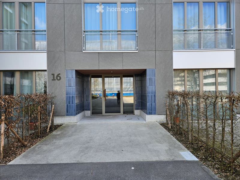 Modern building entrance with large glass doors, numbered '16', and small landscaped area with young trees and shrubs.
