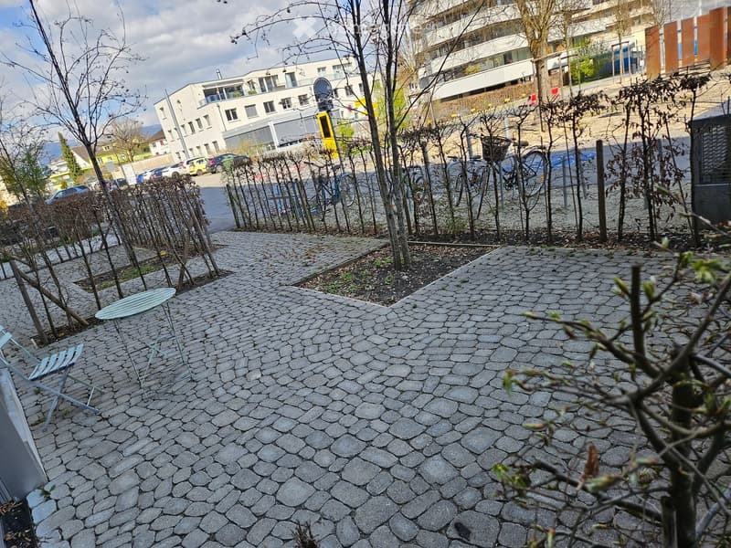 A paved outdoor area with a small table and chairs, surrounded by young trees and a parking area with cars in the background.