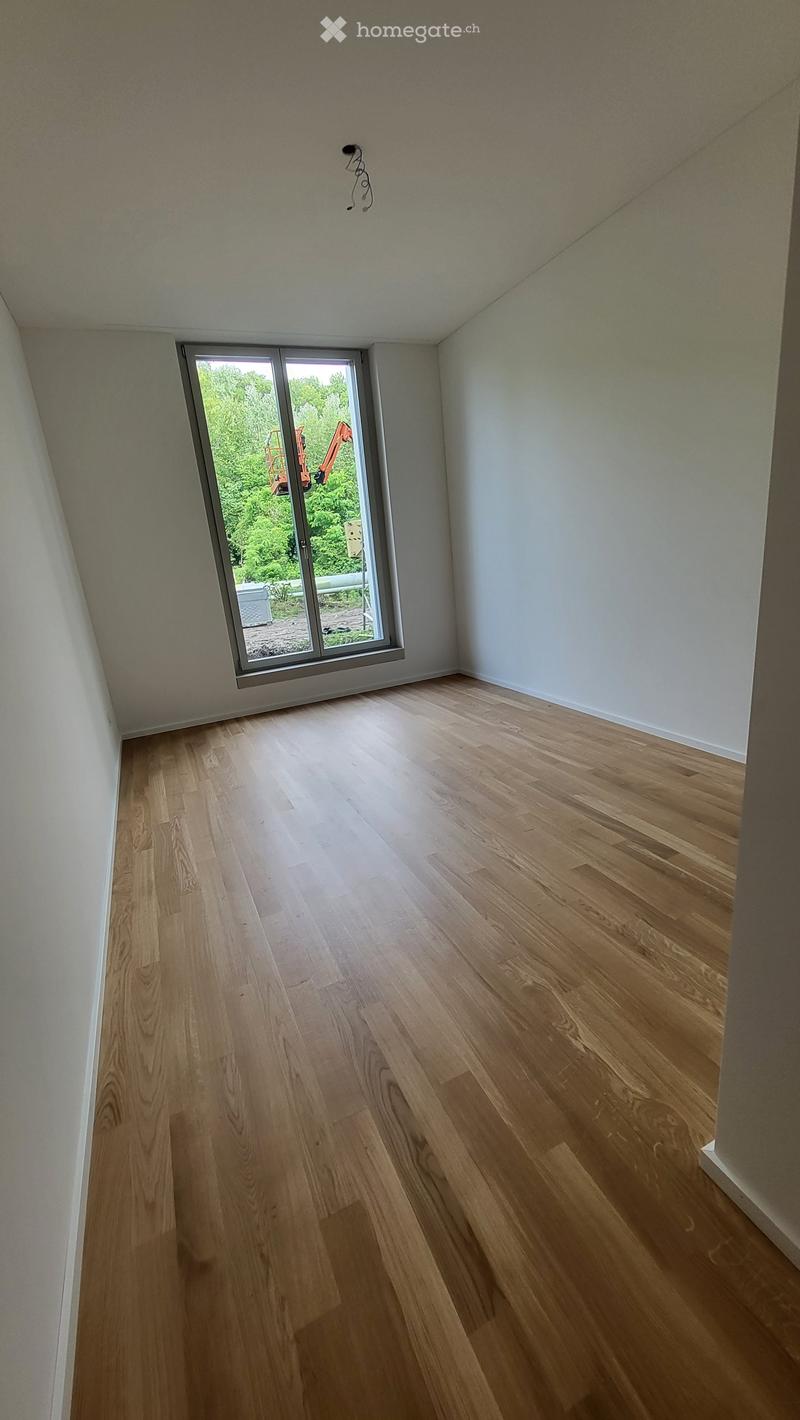 Empty room with light wooden flooring and white walls, featuring a large window with a view of greenery and construction equipment outside.