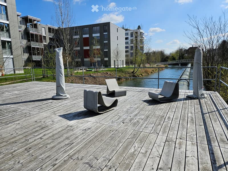 Wooden deck with modern chairs, overlooking a river and modern buildings.