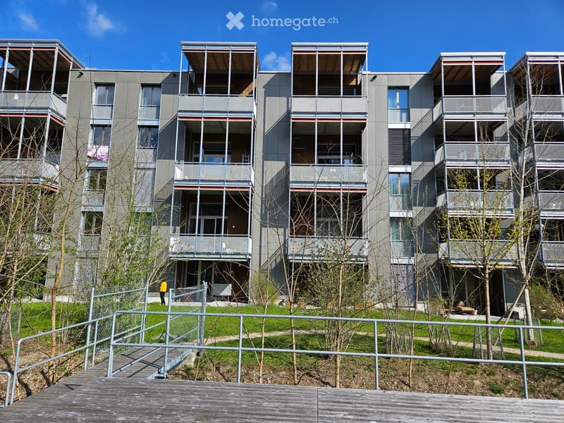 Modern multi-story residential building with grey facade, large windows, and balconies. The building is surrounded by a landscaped area with grass, trees, and a wooden walkway.