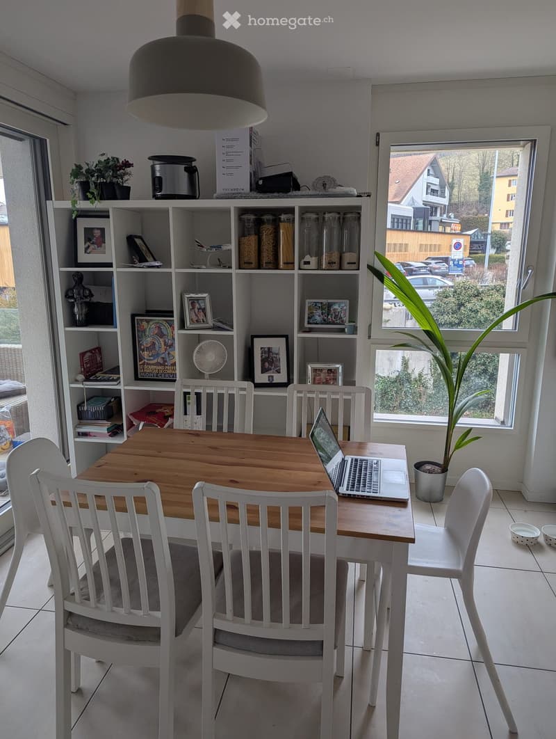 Bright, modern dining area with a wooden table, white chairs, laptop, and a shelf with jars, books, and decorative items. A potted plant is near the window, and the room has large windows with a view of the street.