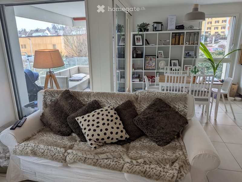Modern living room with a white sofa, fur throw, patterned pillow, dining table with chairs, bookshelf, and large windows with a view.