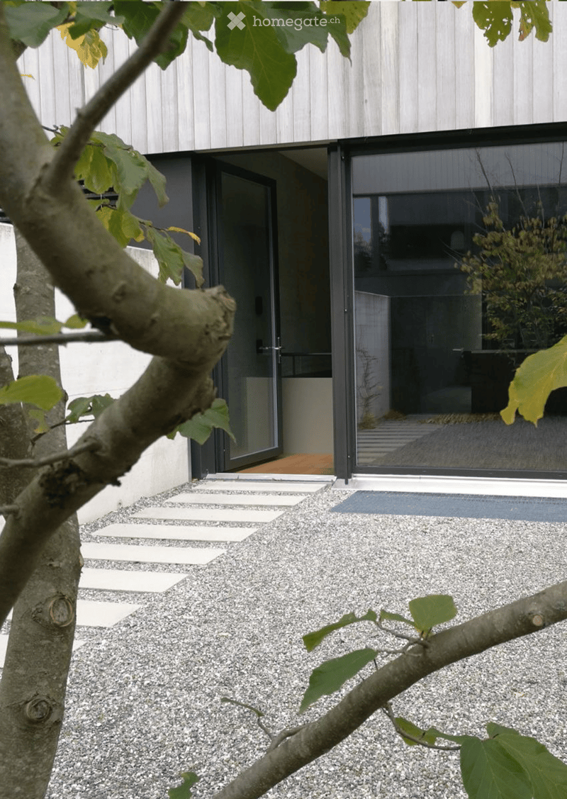 Modern outdoor area with gravel ground, stepping stones, and a tree with lush green leaves. The entrance features large glass doors leading into the building.