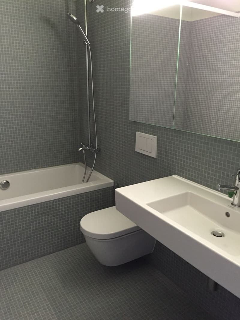 Modern bathroom with dark grey tiled walls and floor, featuring a bathtub, toilet, sink, and showerhead.