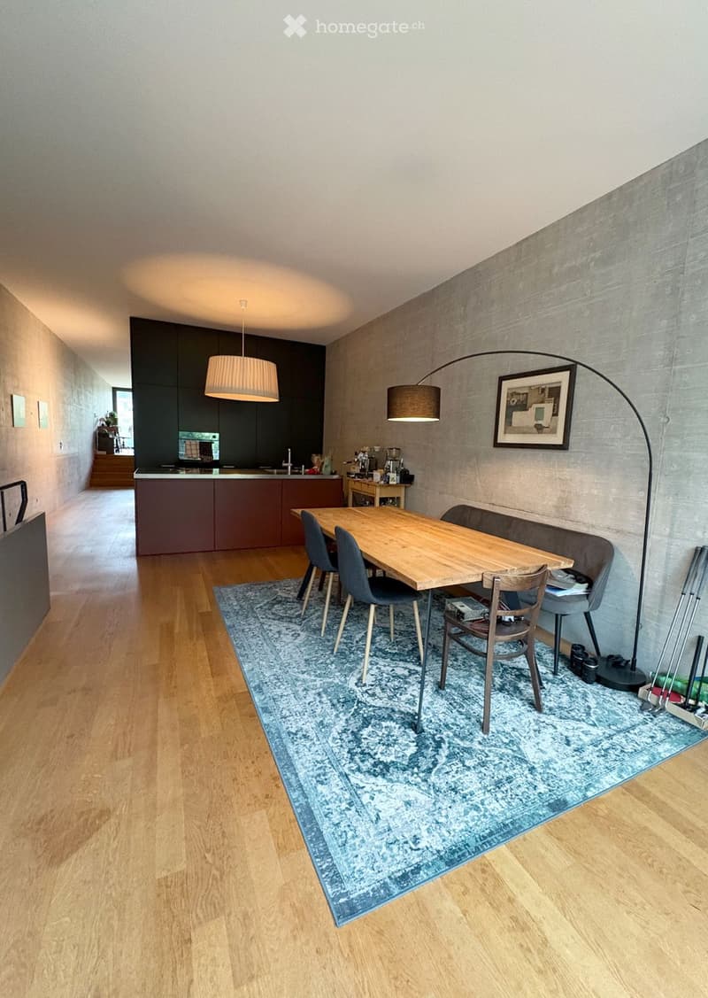 Dining area with a wooden table, four chairs, a sofa, a pendant light, a floor lamp, a framed picture, and a kitchen area with cabinets and a coffee machine.